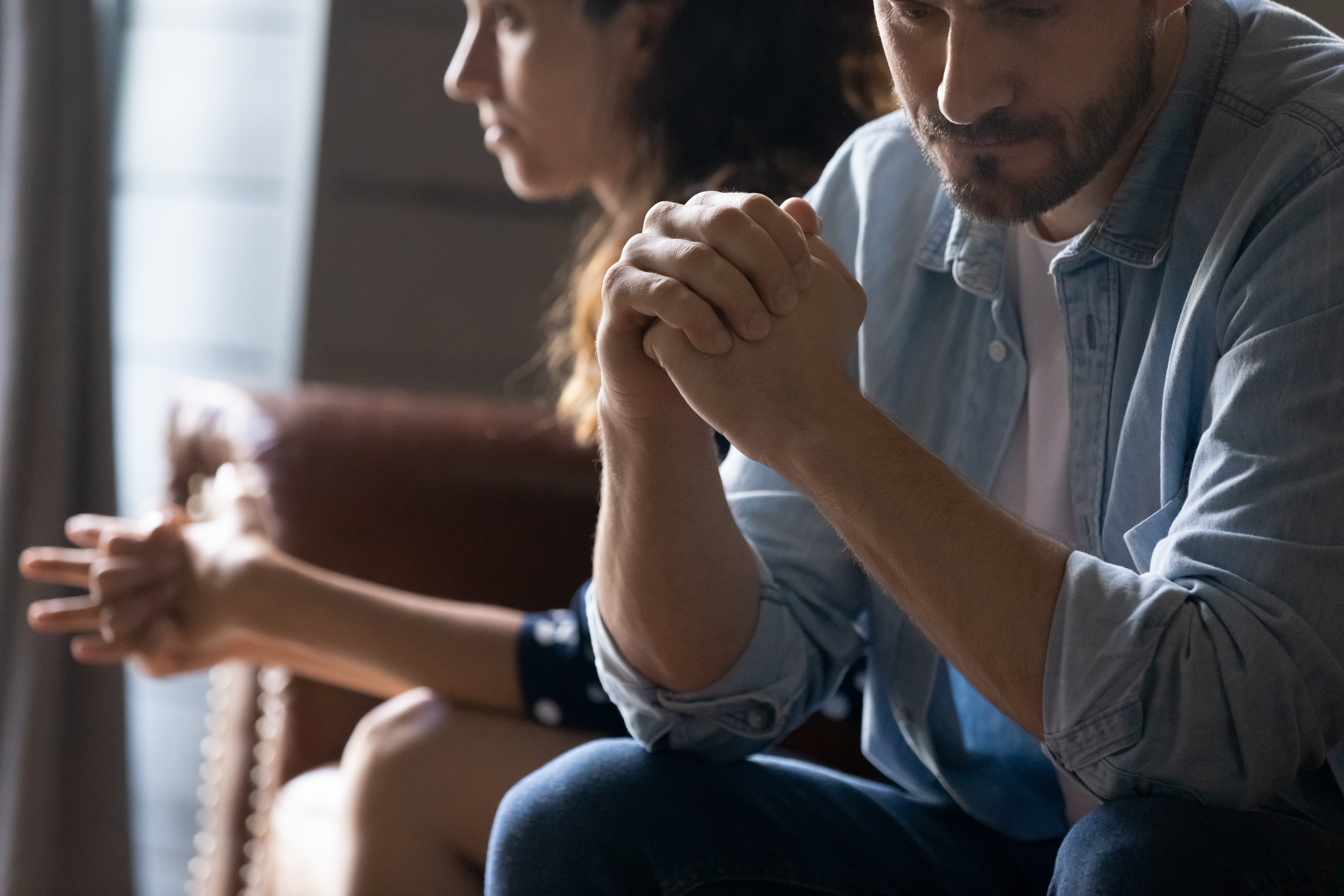 Close-up of a frustrated husband with clasped hands sitting on a couch apart from his wife after an argument, illustrating marriage conflict and the need for leading marriage advice and conflict resolution tools.