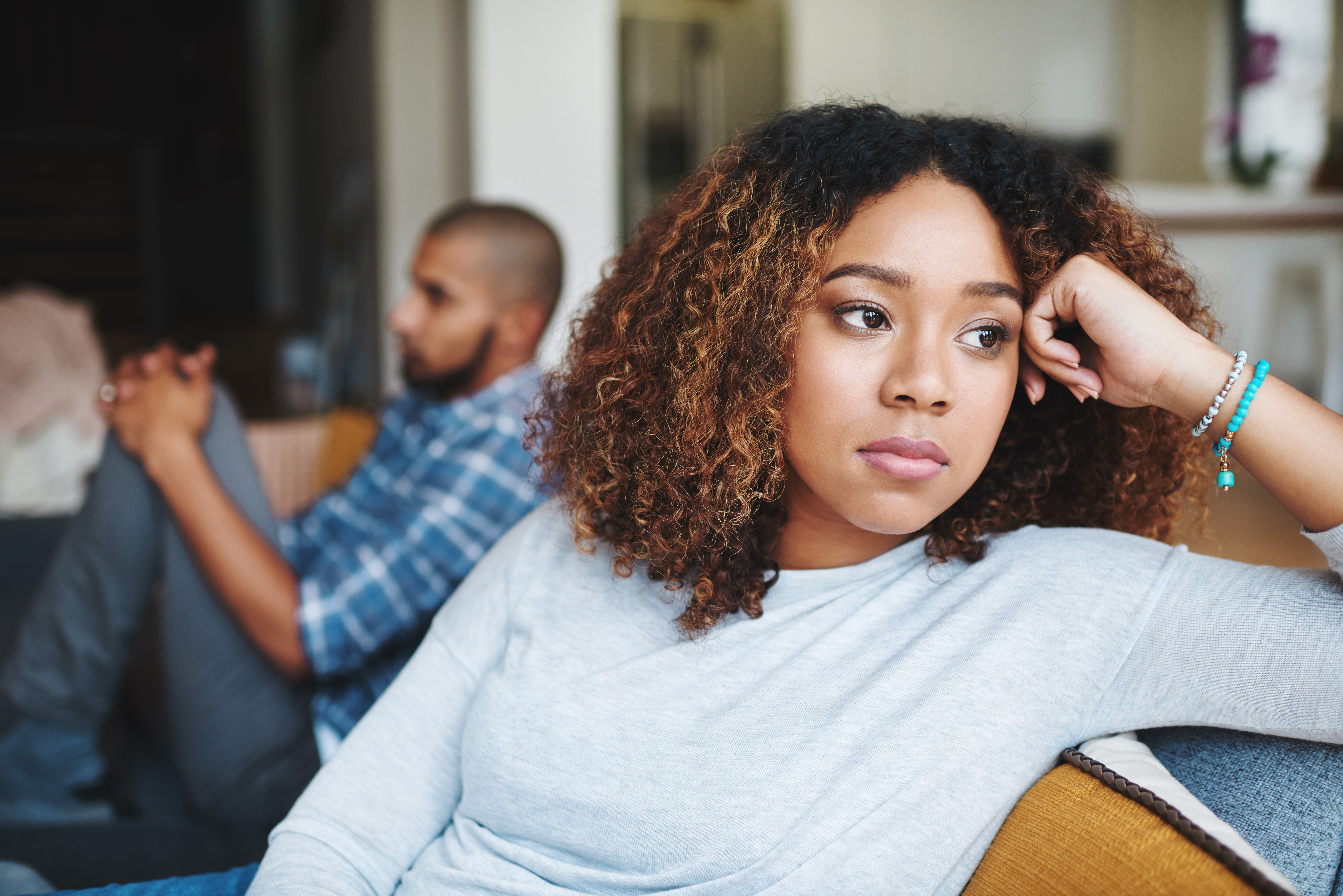 A sad, disconnected wife leaning on her hand on the sofa while her husband sits in the background facing away, representing communication breakdown and the path to restoring a healthy and strong Christian marriage.