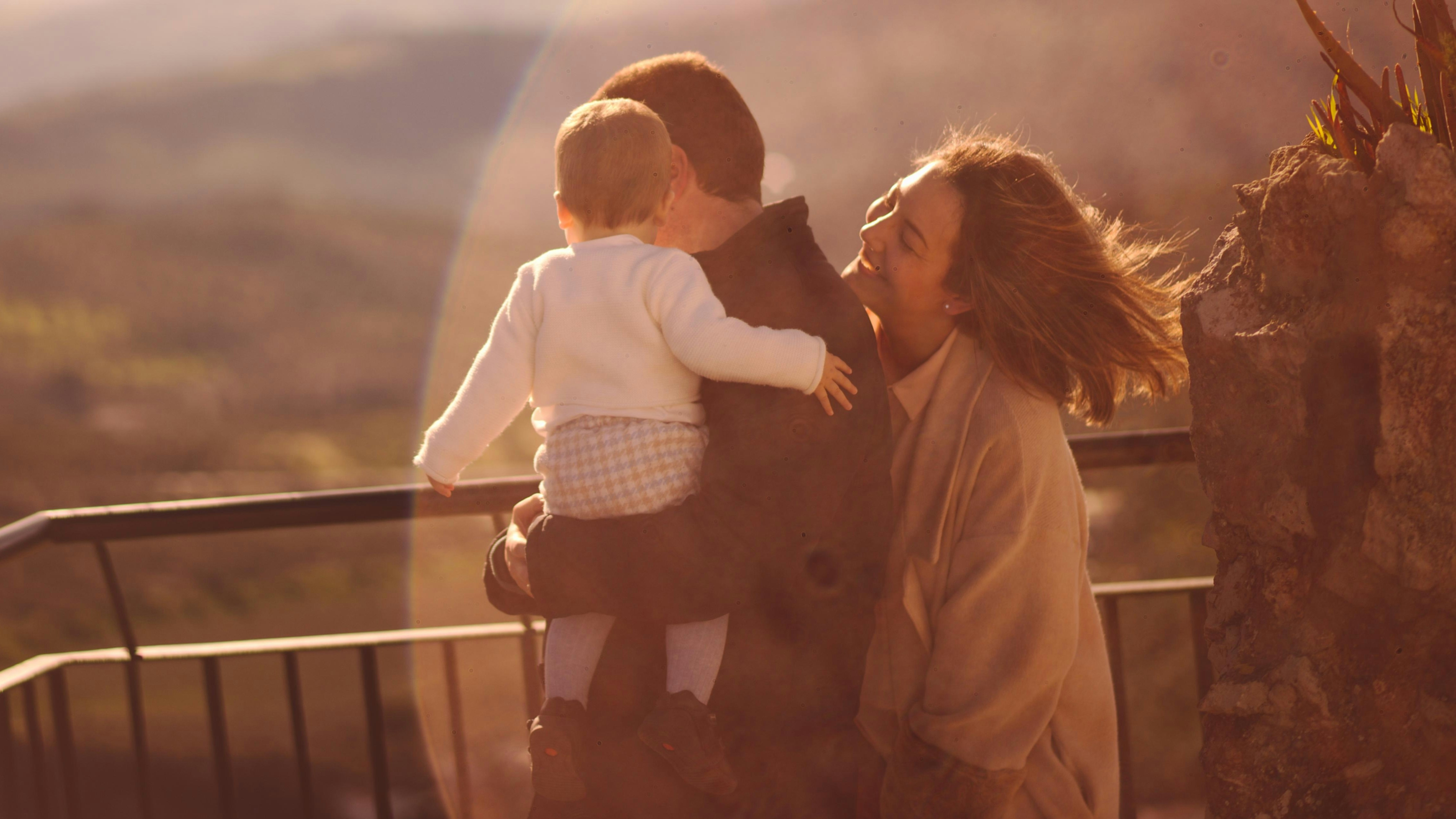 A happy mother and father standing outdoors at sunset holding their toddler, showcasing a thriving family dynamic built on a strong and successful marriage.