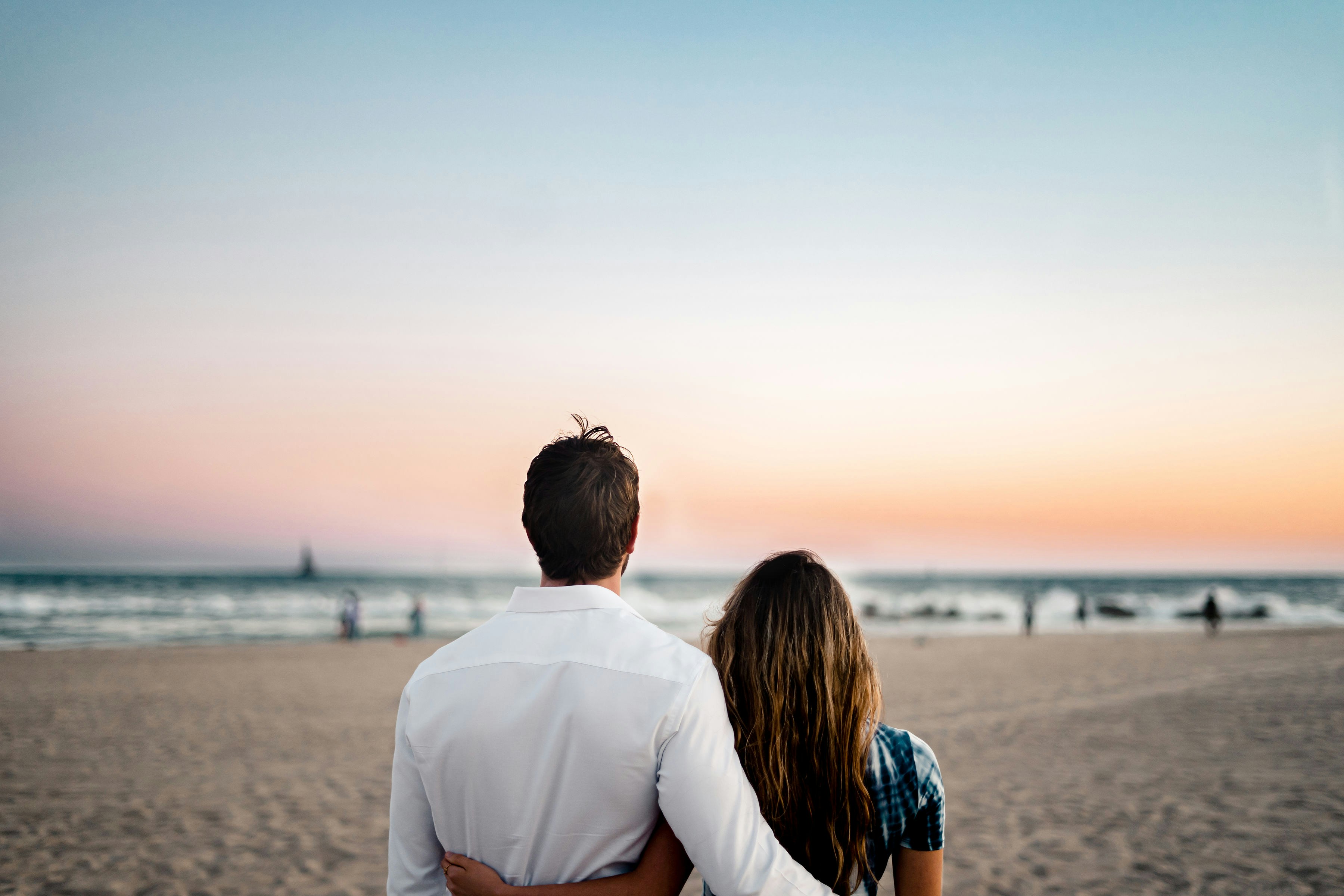 Rear view of a husband and wife standing close together on the beach, looking out at the ocean horizon during a peaceful sunset, representing a unified vision and lifelong commitment in marriage.
