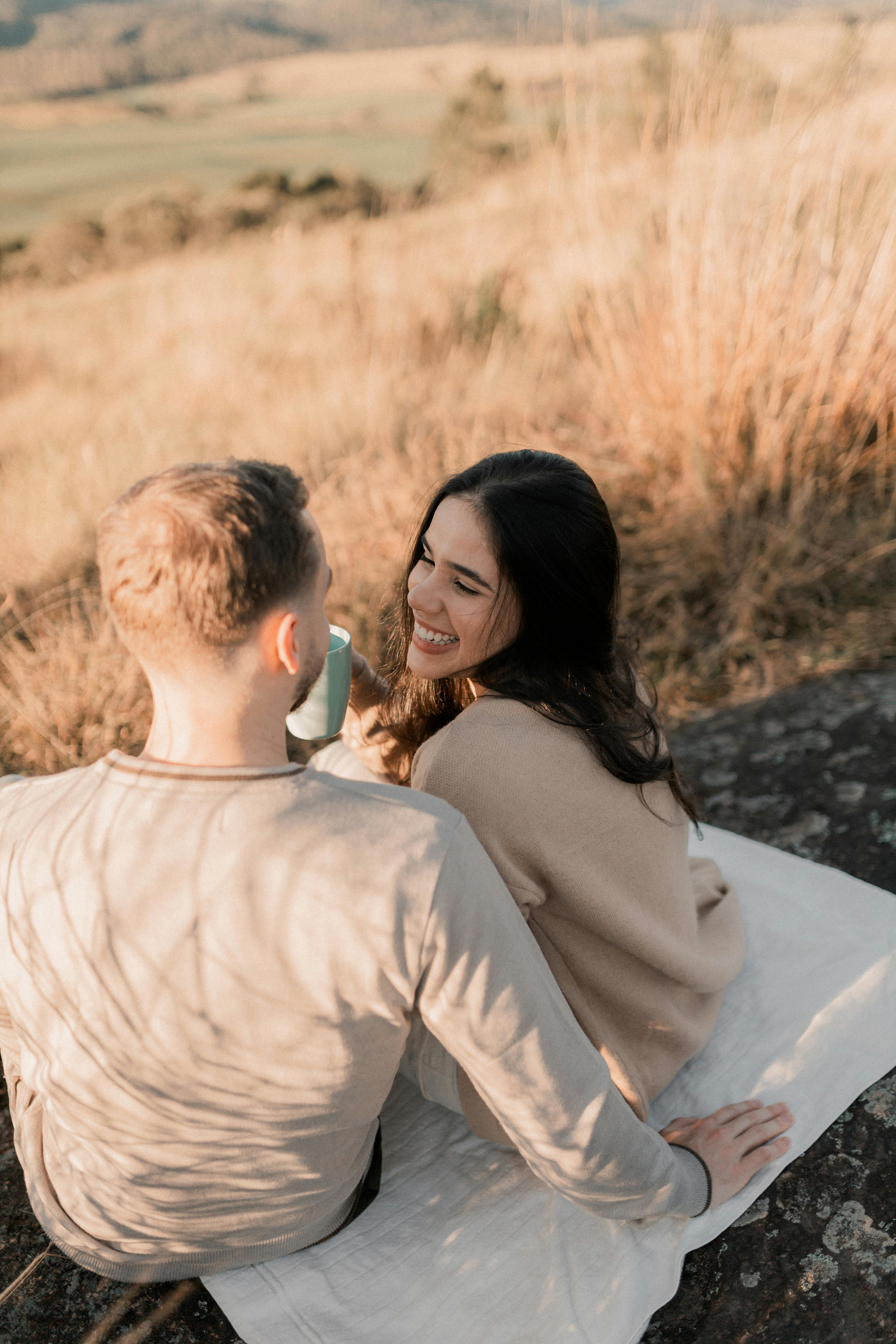 A smiling young husband and wife sitting on a picnic blanket outdoors, sharing a conversation over coffee to illustrate healthy communication and quality time in marriage.