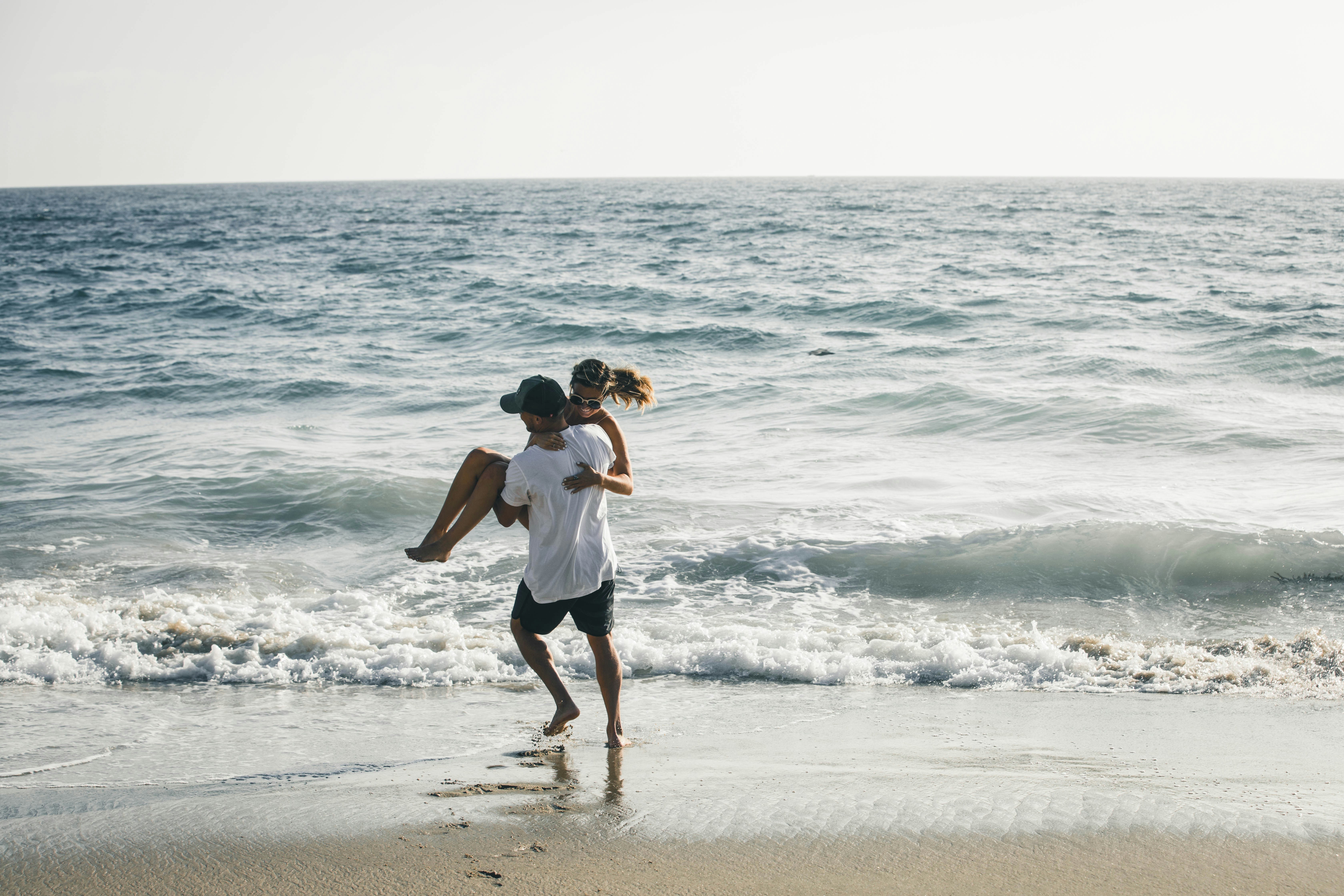 A husband playfully carrying his wife on the beach as ocean waves crash around them, illustrating a fun, thriving relationship and leading marriage advice for keeping romance alive.